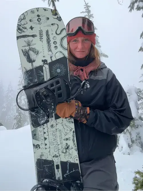 Burton Rewind 2026-2027 On Snow Demo | Snowboarding Profiles Our Snowboard Tester Laura wearing red goggles and a dark jacket holds the Burton Rewind 2027 snowboard at the resort during her testing of it.